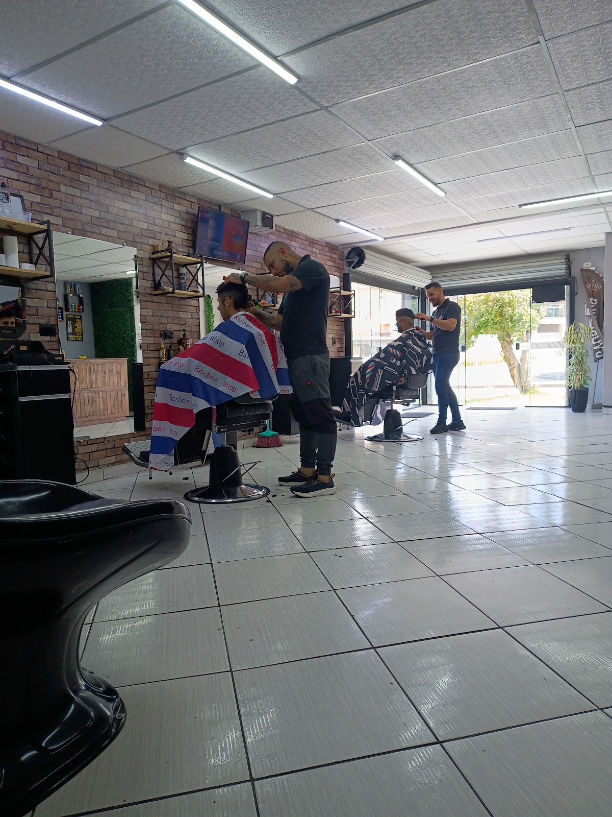 Dois barbeiros cortando o cabelo de clientes ao mesmo tempo, com parede de tijolo aparente ao fundo